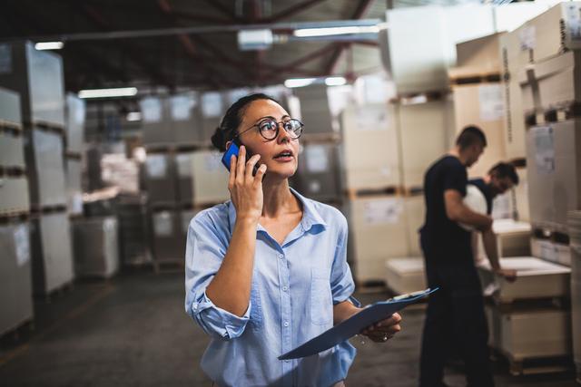 Woman in a warehouse holding a clipboard and talking on a phone while colleagues check inventory behind her