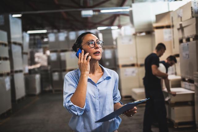 Woman in a warehouse holding a clipboard and talking on a phone while colleagues check inventory behind her