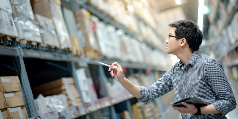 Man in glasses and plaid shirt points with a stylus while holding a tablet among tall warehouse shelves
