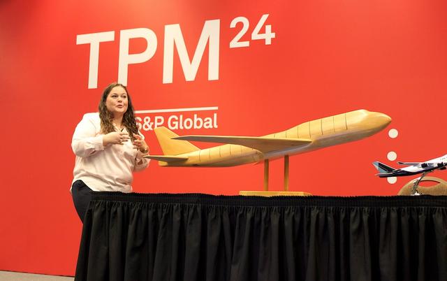 Woman speaks on stage beside a wooden airplane model against a red 'TPM24 S&P Global' backdrop.