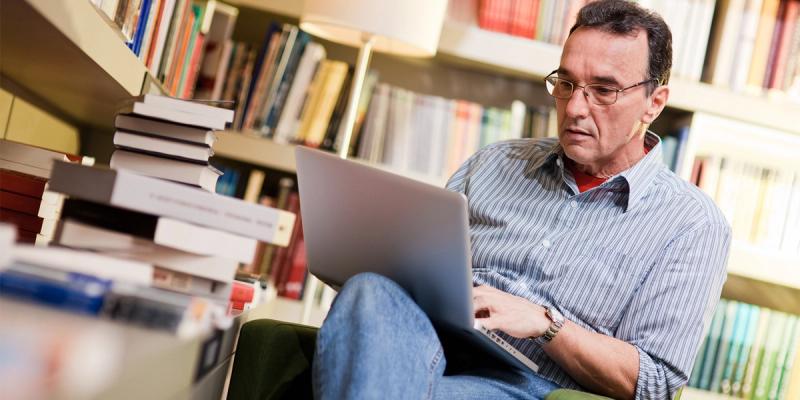 Man in glasses and a striped shirt works on a laptop while seated beside a tall stack of books and library shelves.