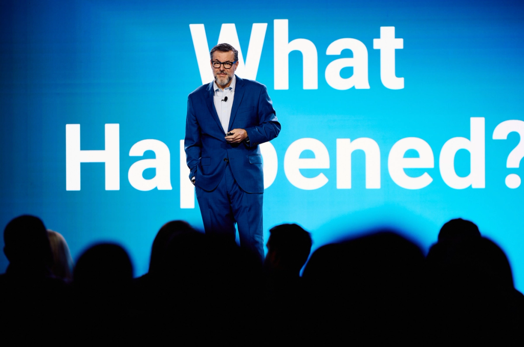 Man in blue suit on stage before a large screen reading What Happened?, audience silhouetted in foreground