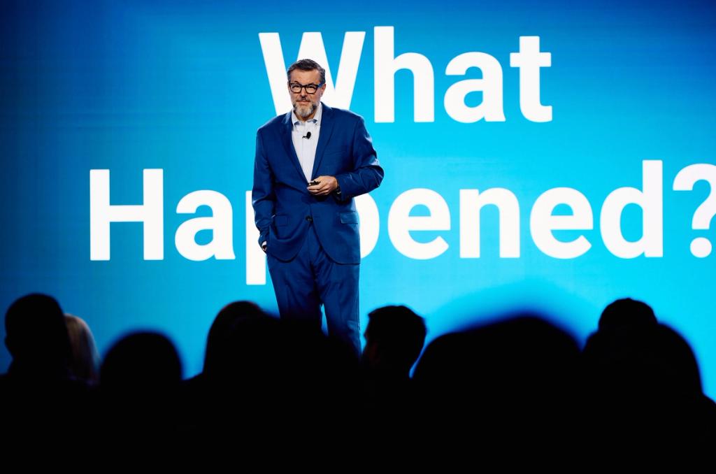 Man in blue suit on stage before a large screen reading What Happened?, audience silhouetted in foreground
