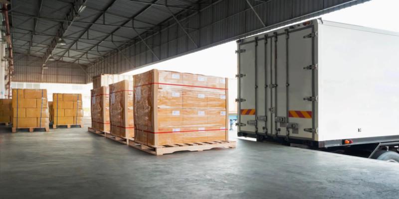 Shrink-wrapped pallets of boxes on a warehouse floor beside a white box truck at a loading dock