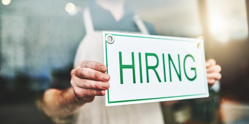 Aproned worker holds a white sign reading HIRING in green letters against a blurred storefront