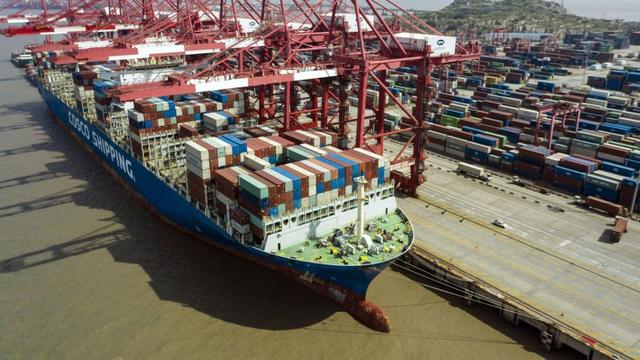 Aerial view of a COSCO Shipping container vessel docked at a port beneath red cranes, surrounded by stacked shipping containers