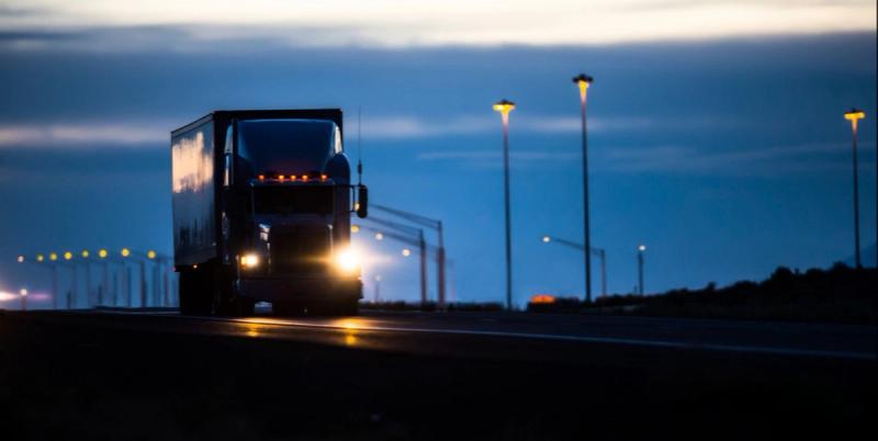 Semi-truck driving on a highway at dusk with headlights on and streetlights glowing.