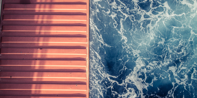 Aerial view of an orange container ship hull cutting through churning blue ocean water.