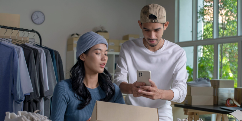 Young man and woman in a small ecommerce warehouse looking at a smartphone, with clothing racks and shipping boxes