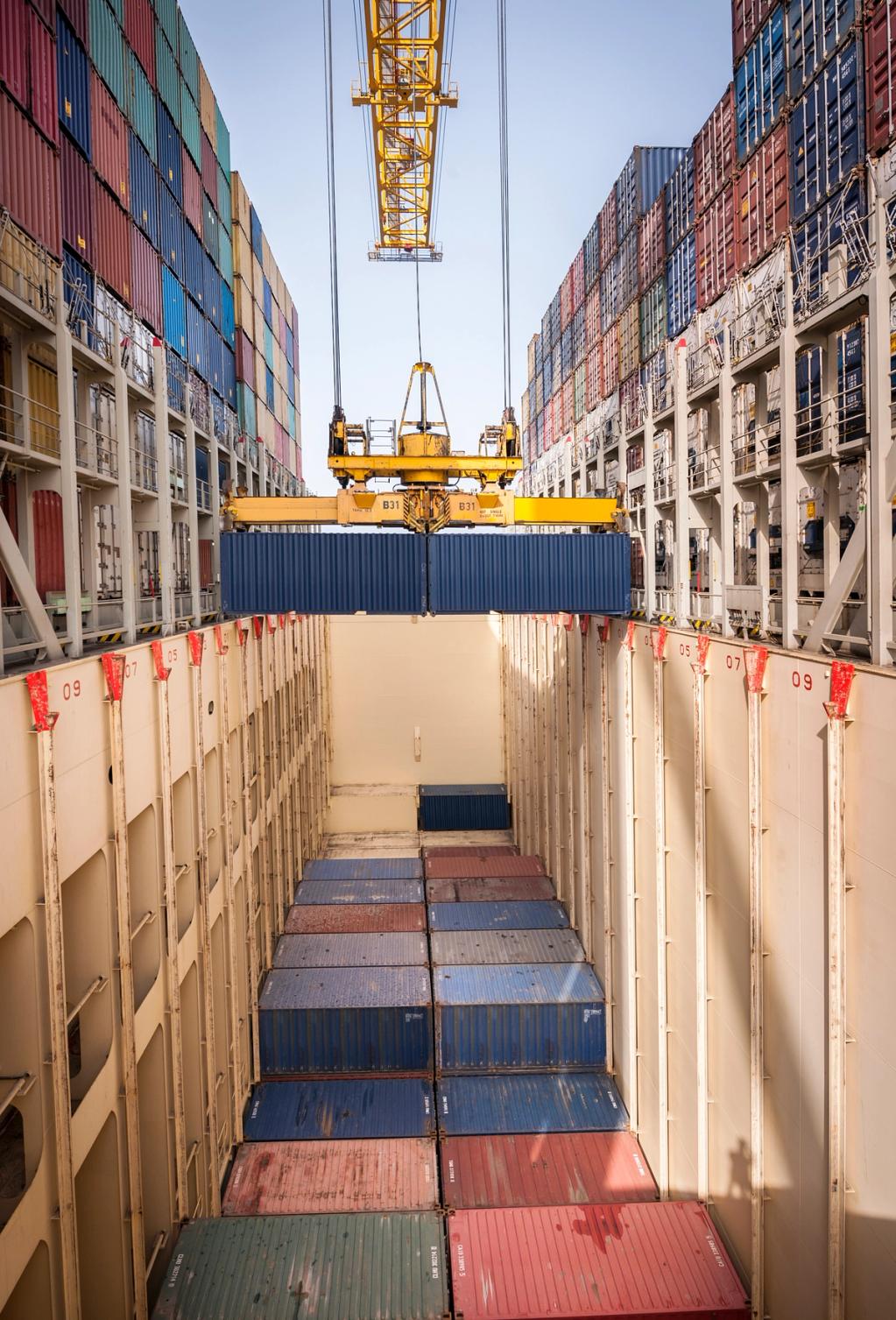 Looking down a container ship hold as a gantry crane lowers a blue container onto stacked rows of boxes
