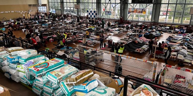 Indoor relief center with rows of cots and stacks of diapers and supplies for refugees