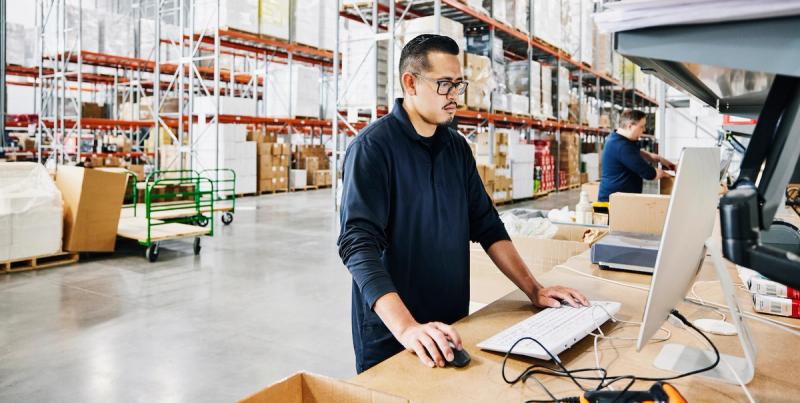 Man in glasses working at a computer at a counter inside a warehouse with tall stocked shelves