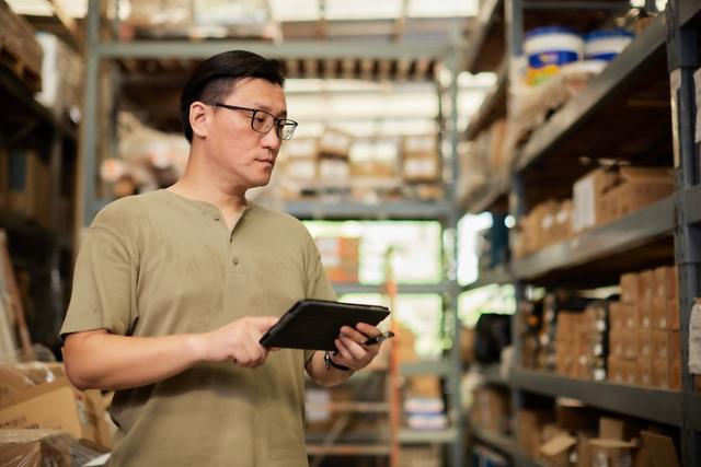 Man in glasses checking inventory on a tablet beside warehouse shelves stocked with boxed parts