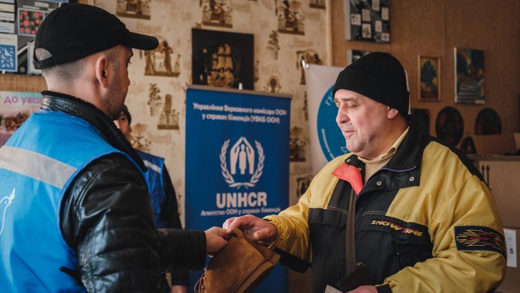 Aid worker in a blue vest hands an item to a man at a UNHCR distribution point with a UNHCR banner