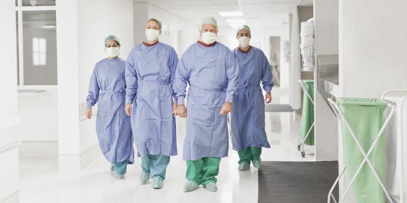 Four masked medical staff in blue surgical gowns and green scrubs walk down a bright hospital corridor