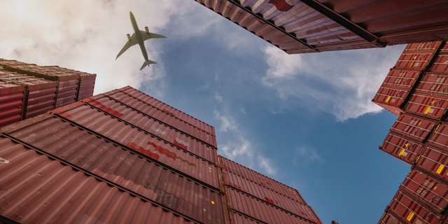 Upward view of stacked shipping containers framing a bright sky with an airliner passing overhead