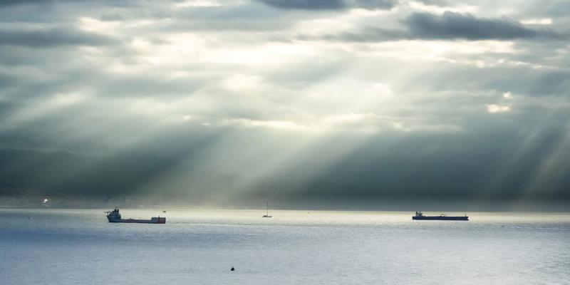 Sun rays break through storm clouds over calm water with cargo ships and a sailboat in the distance