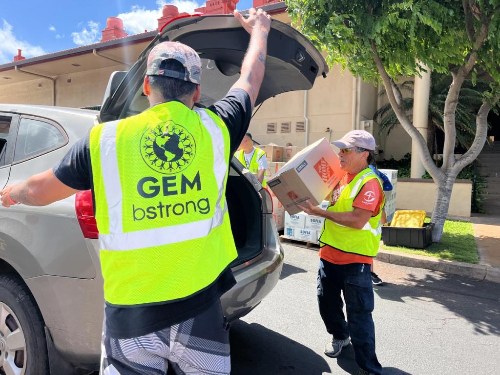 Volunteers in GEM bstrong and orange vests loading aid boxes into a car trunk outside a building