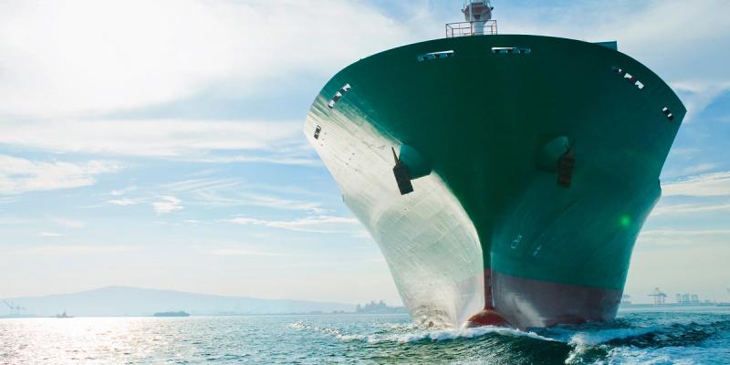 Bow of a green and white container ship cutting through calm sea water near a port