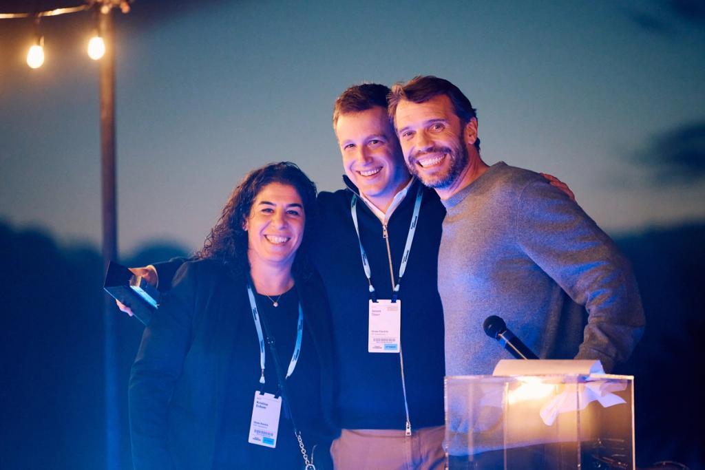 Three smiling people pose together holding an award on a stage at a dusk outdoor event