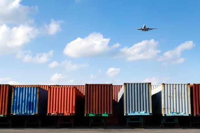 Row of stacked shipping containers on rail cars with a passenger jet flying overhead against a blue sky