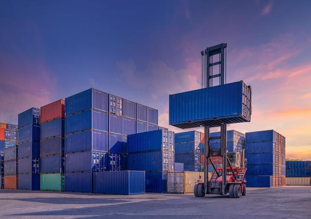 Reach stacker lifts a blue shipping container above stacks of containers in a port yard at sunset