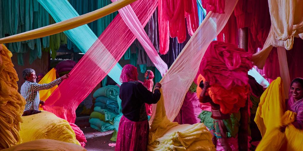 Workers handling sheets of brightly dyed fabric hung overhead in a colorful textile dyeing workshop