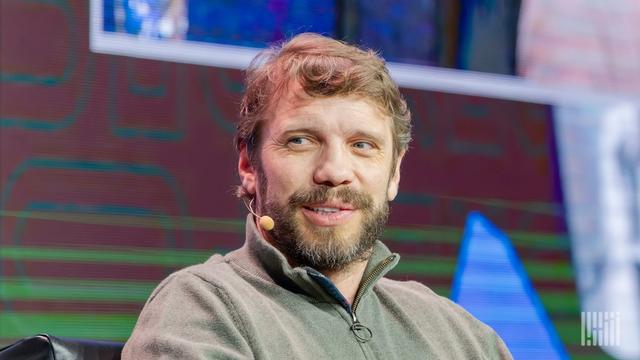 Bearded man in a gray quarter-zip sweater wearing a headset mic, seated on stage smiling before a colorful event backdrop