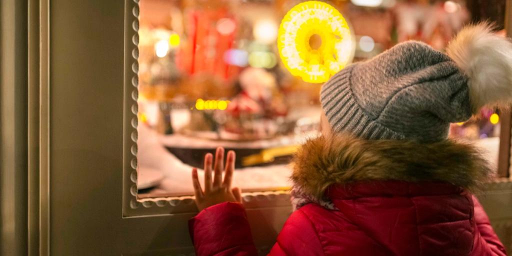 Child in red coat and gray pompom hat presses a hand to a festive holiday shop window