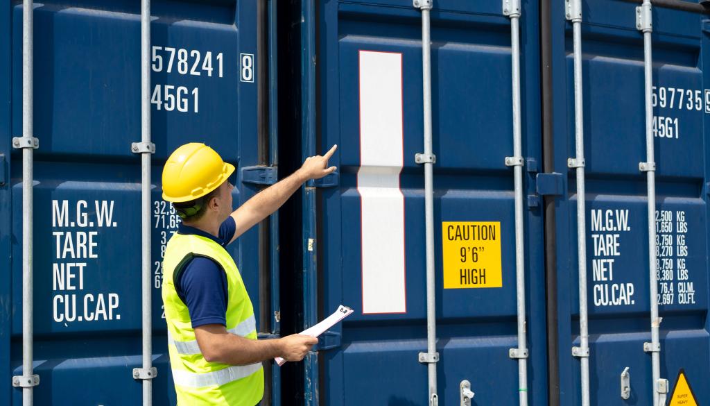 Worker in a hard hat and hi-vis vest inspecting and pointing at stacked blue shipping containers, holding a clipboard.