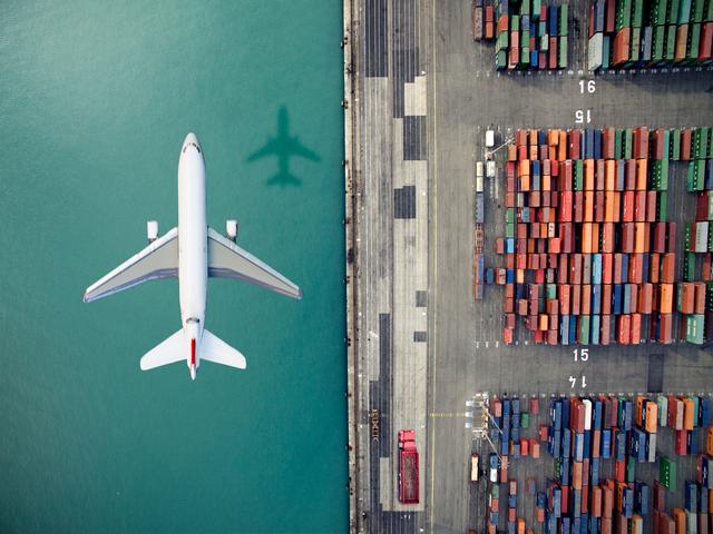 Aerial view of a white airliner flying over teal water beside a port stacked with colorful shipping containers