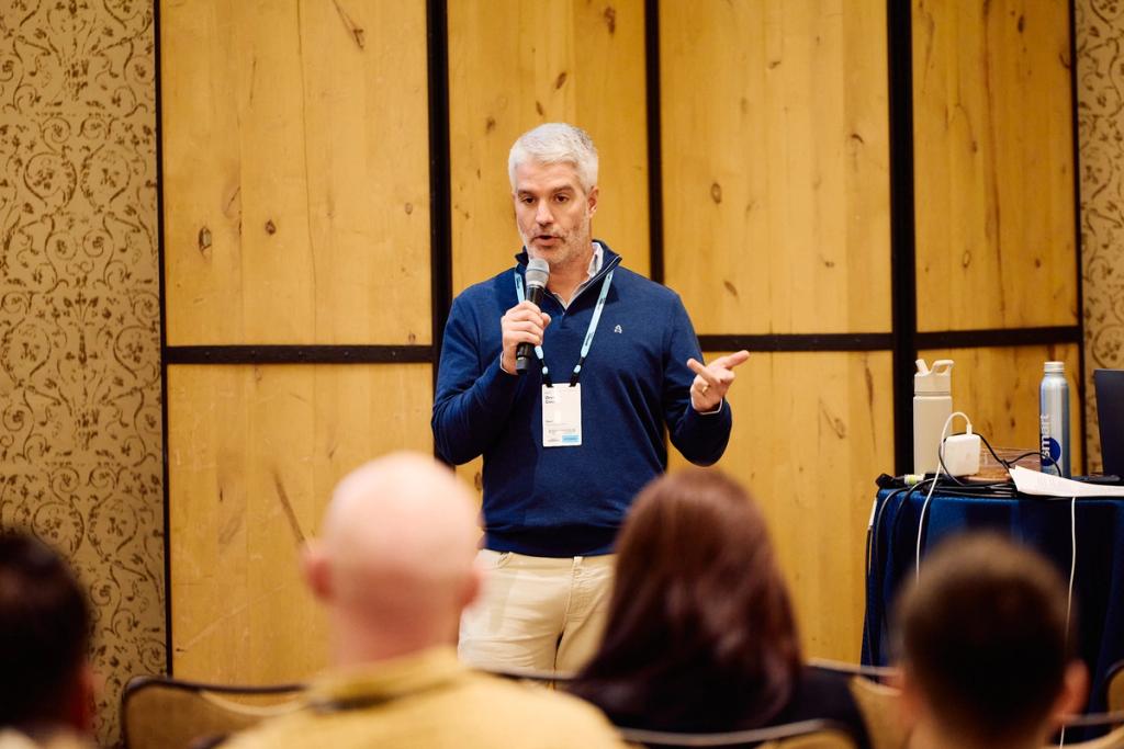 Man in navy sweater speaks into a microphone to a seated audience at a wood-paneled conference session.