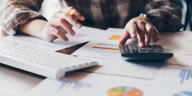 Person in plaid shirt using a calculator over financial charts beside a keyboard