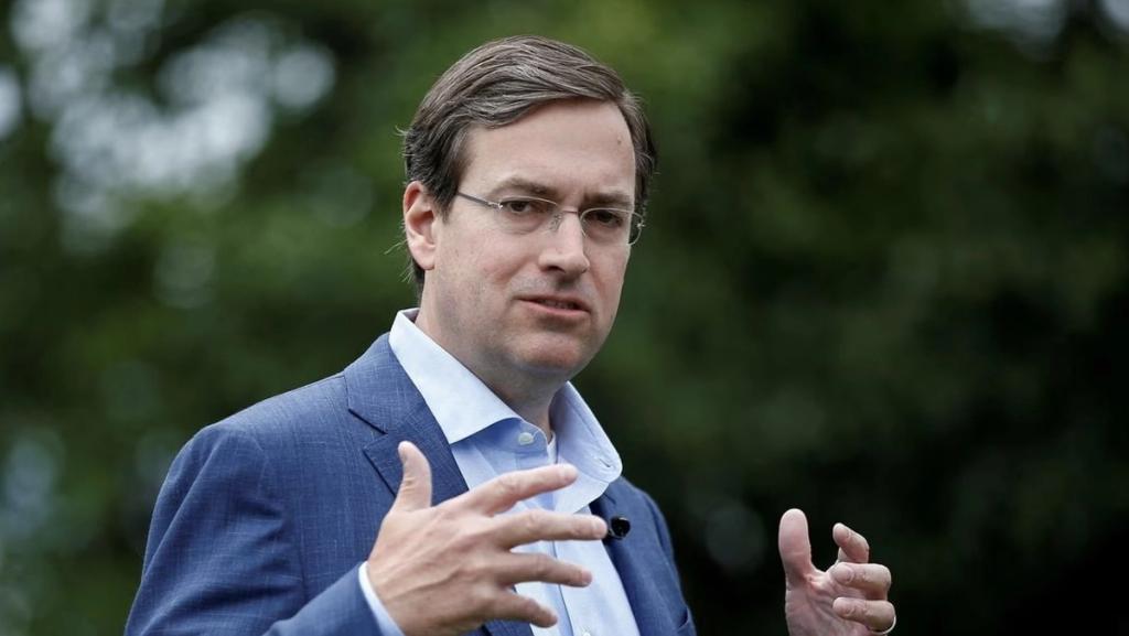 Man in a blue blazer and light shirt with glasses gesturing while speaking outdoors against a leafy green background