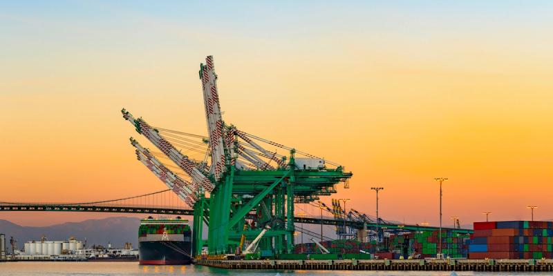 Green gantry cranes and a container ship at a port at sunset, with a suspension bridge and stacked containers
