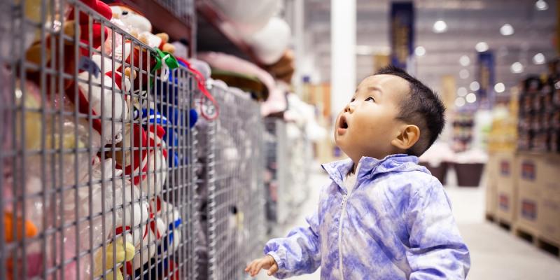 Young child in a blue jacket gazing up at plush toys stacked in a store aisle