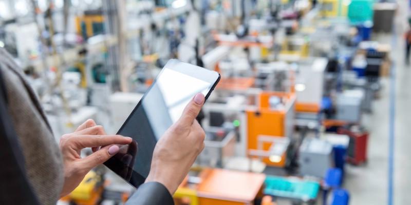 Hands use a tablet overlooking a busy factory floor with machinery below