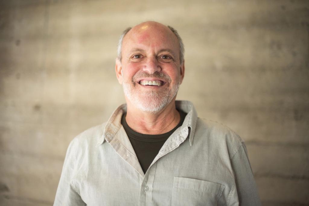 Smiling bald man with grey beard in a light shirt against a concrete wall