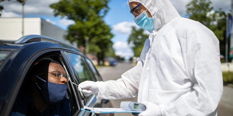Health worker in white PPE and mask swabs a masked person seated in a car at a drive-through testing site