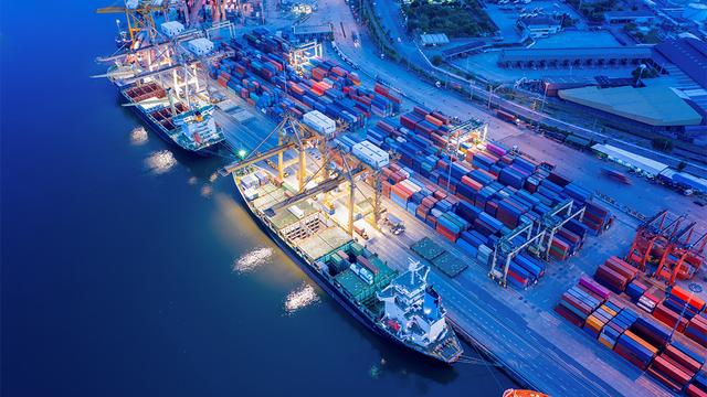Aerial view at dusk of a busy container port with cargo ships, gantry cranes, and rows of stacked shipping containers