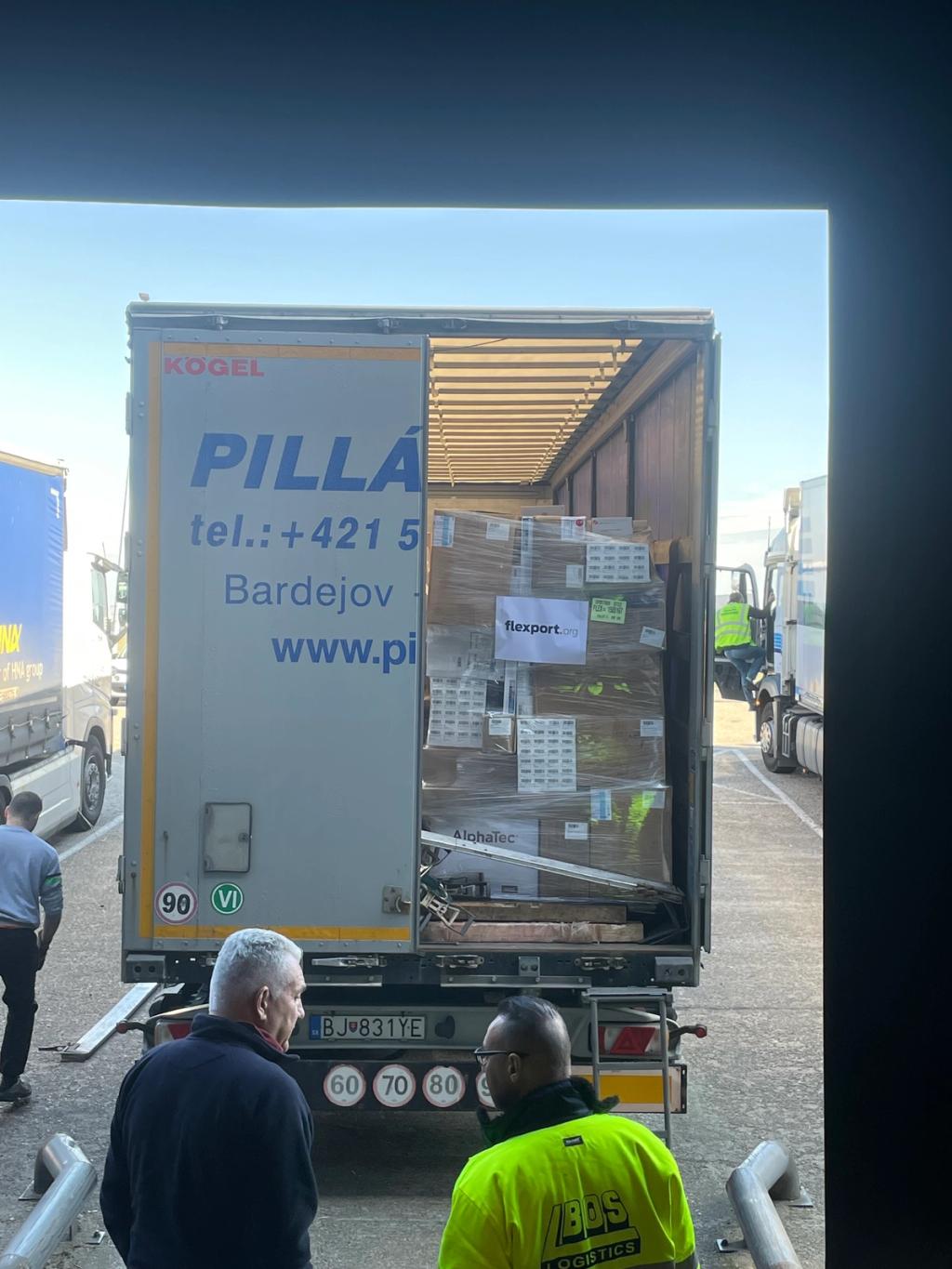 Two men view the open rear of a Pilla truck trailer loaded with palletized boxes labeled flexport.org