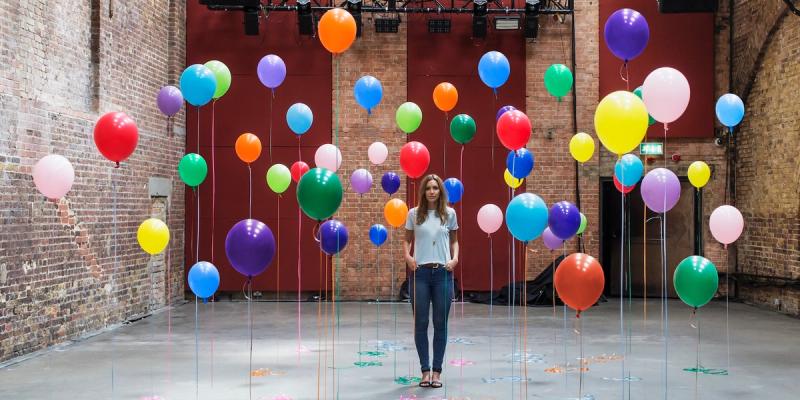 Woman standing in a brick warehouse surrounded by dozens of colorful balloons rising on strings from the floor