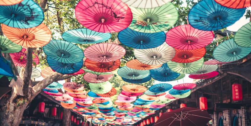 Canopy of colorful paper parasols strung over a tree-lined Chinese street with red lanterns