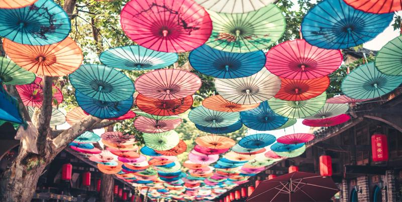 Canopy of colorful paper parasols strung over a tree-lined Chinese street with red lanterns