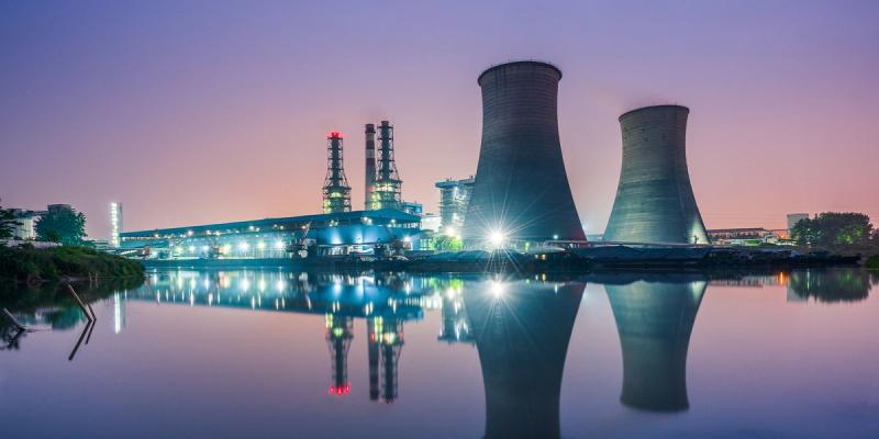 Power plant with cooling towers and smokestacks lit at dusk, reflected in calm water