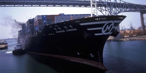The container ship Hanjin London, bow forward and stacked with containers, moves through a port channel under a bridge.