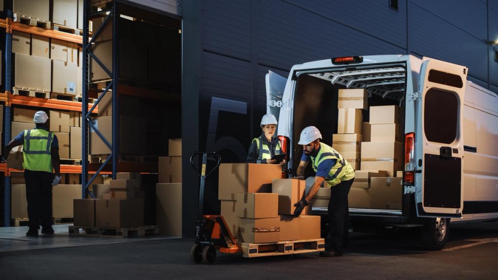 Two workers in hard hats and hi-vis vests loading boxes from a pallet jack into a delivery van at a dimly lit warehouse loading bay