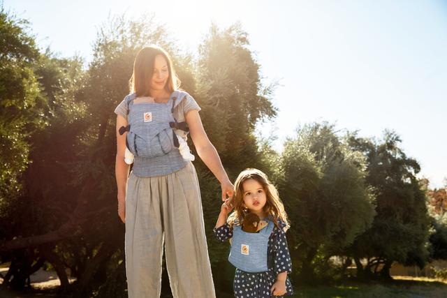 Woman wearing an Ergobaby carrier holds hands with a toddler in a sunlit garden