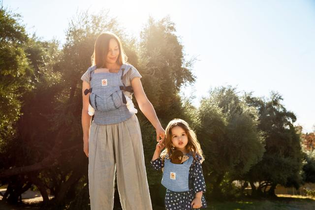 Woman wearing an Ergobaby carrier holds hands with a toddler in a sunlit garden