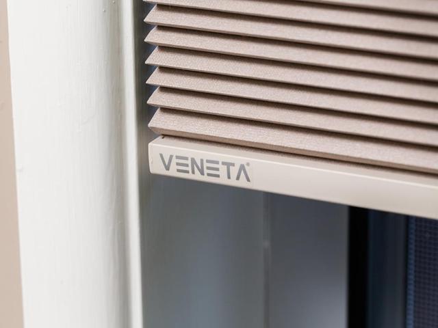 Close-up of pleated beige Veneta window blinds with the brand logo on the bottom rail